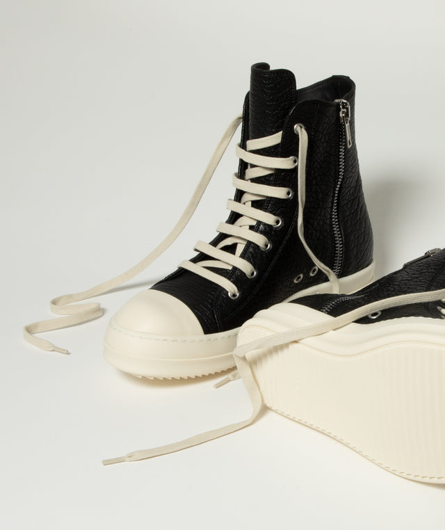 A pair of Rick Owens black high-top sneakers with textured leather uppers, cream laces, and thick cream platform soles, shown on a light gray studio background. 