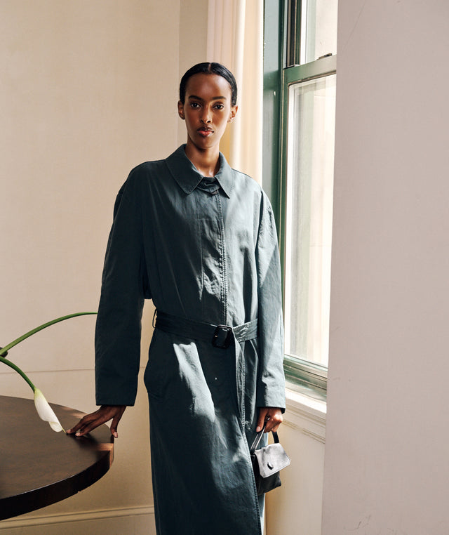 A model stands by a window wearing a dark slate blue-gray long-sleeve collared shirt dress with a matching belt by The Row. She is holding a small button handbag by The Row. 