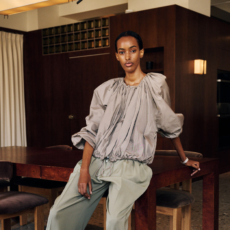 A model wearing a light gray oversized bubble-sleeve coat by Toteme and sage green wide-leg trousers, leaning against a dark wooden table in a modern interior setting.
