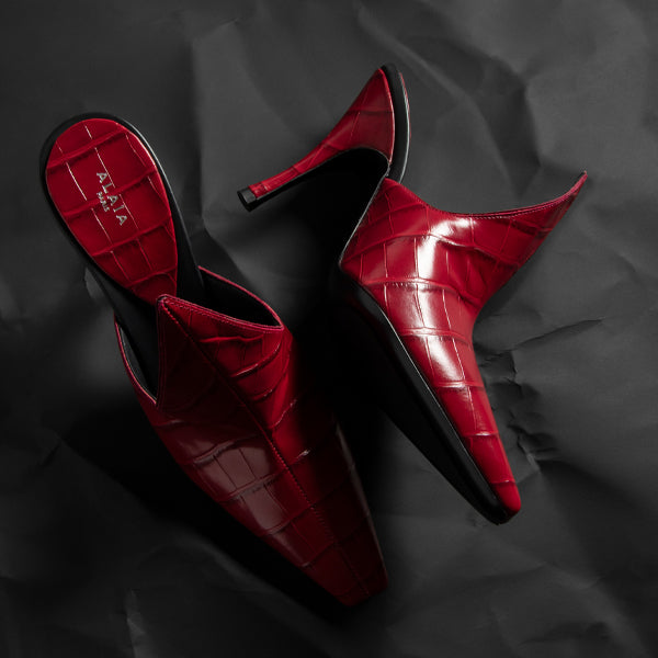 ALAIA red pointed-toe mule heels with a glossy crocodile-embossed texture and black mid-height stiletto heels, photographed on a dark crumpled fabric background. 