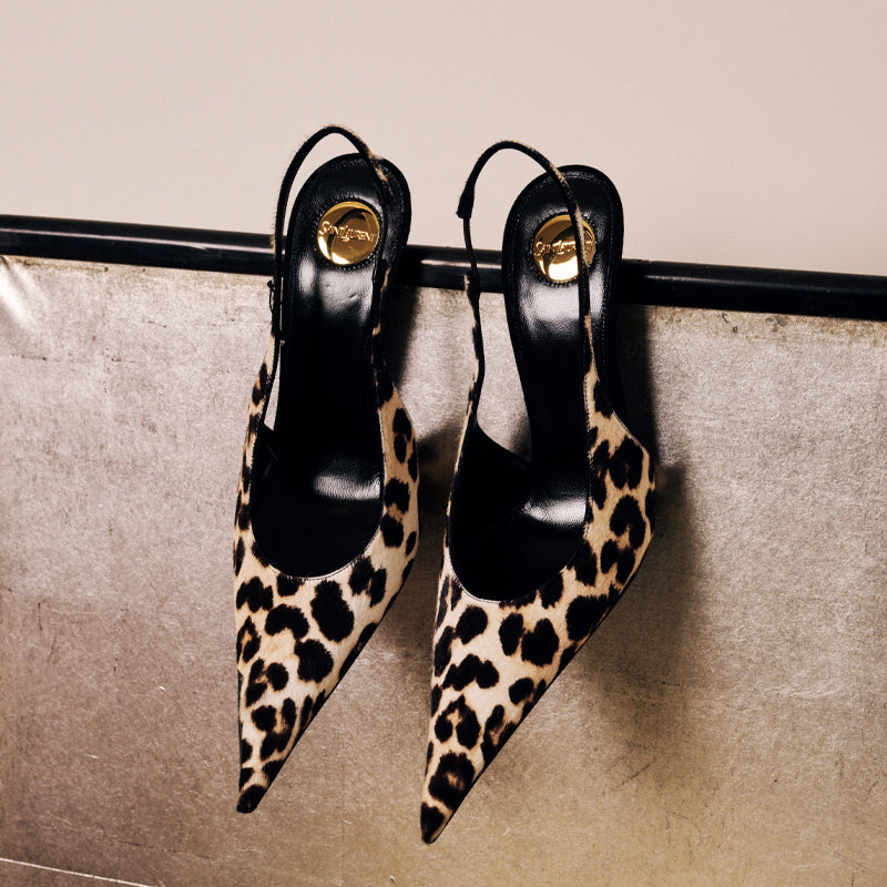 A pair of pointed-toe slingback heels in leopard print with black insoles and gold-tone hardware, hanging from a black rail against a textured beige wall.