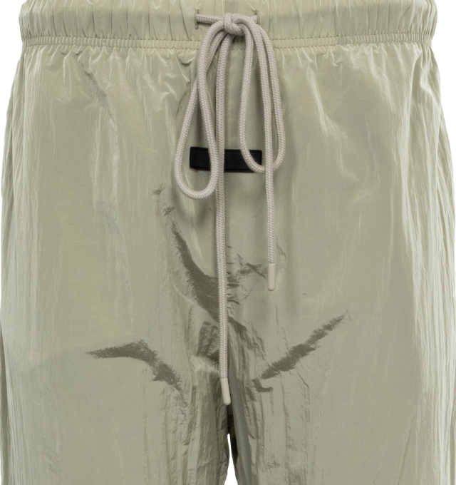 Image 4 of 4 - GREEN - FEAR OF GOD ESSENTIALS Crinkle Nylon Trackpants featuring an encased elastic waistband with elongated drawstrings, side seam pockets, an elastic hem with zipper adjustability at the ankle and a rubberized label at the center front. 100% nylon.  