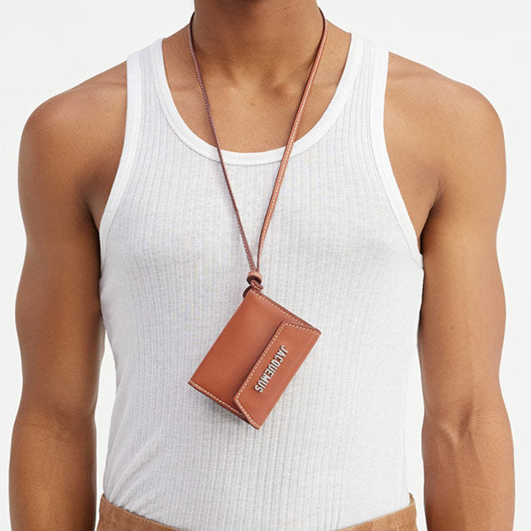 Man's torso wearing Jacquemus brown leather card case hanging by a brown leather strap around the neck, available online.