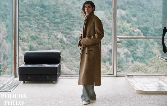 Phoebe Philo campaign - model wearing a long brown coat standing in a modern room with large windows showing a mountain view.
